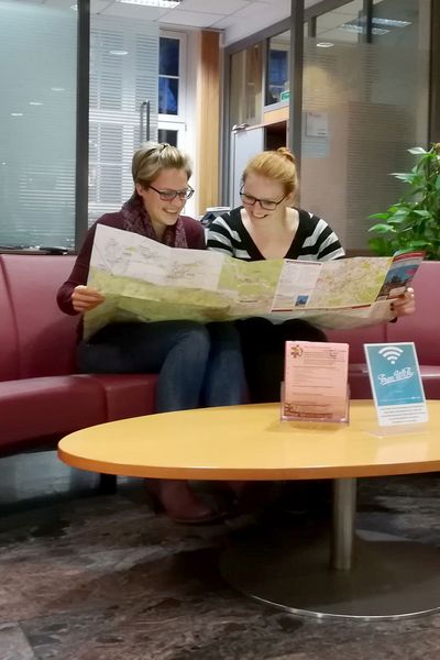 Zwei Personen sitzen auf der roten Couch in der Tourist-Information Wernigerode und schauen in eine Landkarte.