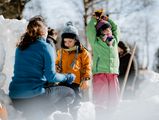 Eine Person sitzt in Hocke mit zwei Kindern im Kurpark Schierke und baut ein Iglo während der Schierker Wintersportwochen.