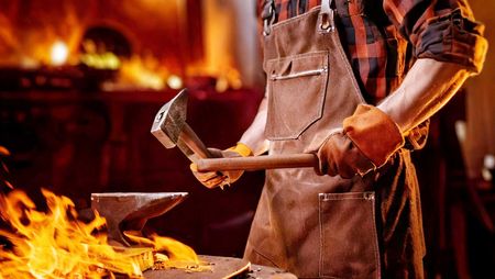 Eine Person steht vor einem Schmiedefeuer und schlägt mit einem Hammer auf einen Amboss